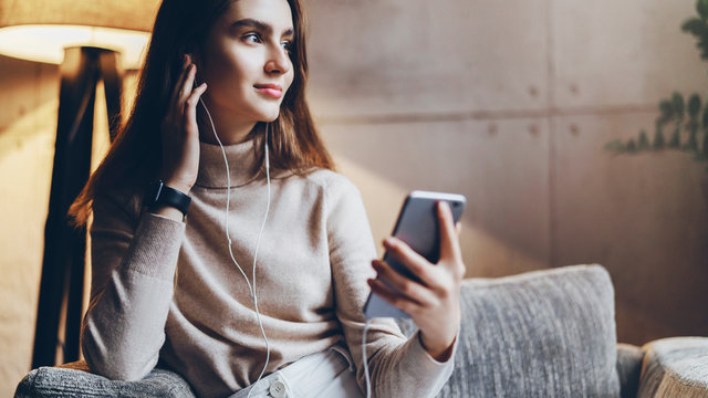 Pretty young woman sitting on sofa and listening to music on smartphone. Girl in earphones streaming songs online in social networks. Student taking language courses remotely