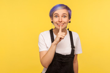 Shh, don't talk! Portrait of positive hipster girl with violet short hair in denim overalls showing silence gesture with fingers on lips, asking to be quiet. isolated on yellow background, studio shot