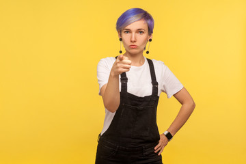 Hey you! Portrait of hipster woman with violet short hair in denim overalls pointing at camera and looking strict rigid, making choice with serious face, selecting you. yellow background, studio shot