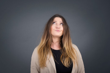 Troublesome puzzled Caucasian woman frowns face, bites lip, raises eyebrows, looks up, has problems, dressed in fashionable clothes, isolated over gray background, has regret look.