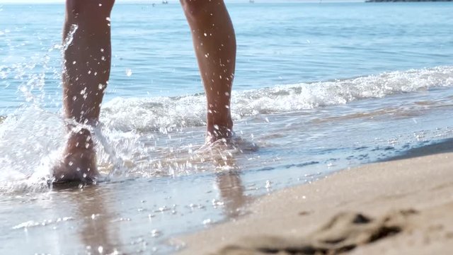 Middle age woman goes along the sandy beach in sunny day. Caucasian woman walking barefoot on sea water. Female legs on ocean waves. Enjoy summer vacation.