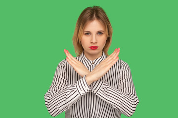 No way! Serious girl with red lips in striped blouse standing with crossed hands, gesturing stop, finish, showing symbol of rejection, body language. indoor studio shot isolated on green background