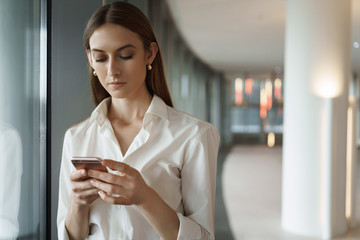 Young elegant lady in white collar shirt checking messages, answering business partner during meeting break, standing in office corridor, using smartphone, texting coworker, rehearsing speech
