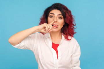 Bad manners, misconduct. Portrait of funny comical hipster woman with fancy red hair picking her nose and sticking out tongue with stupid expression. indoor studio shot isolated on blue background