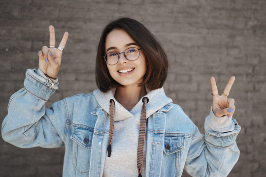 Close-up portrait of queer cute girl in glasses, college student showing kawaii peace gestue and smiling camera, enjoying youth, university carefree life, standing near brick wall on street