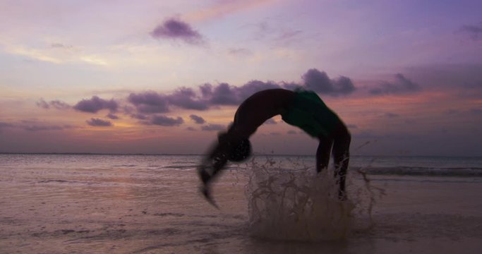 Muscular african capoeira acrobatic athlete is performing tricks on a desert sea water with splash on a sunset. Concept: healthy lifestyle, diet, wellbeing, fitness, proper nutrition, love for sports