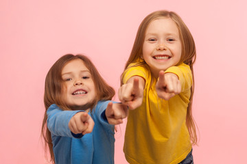 Hey you! Two charming happy little girls pointing to camera, indicating lucky one and smiling, choosing goods in child store, we need this concept. indoor studio shot isolated on pink background