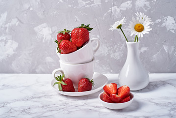 Ripe organic strawberries in white ceramic bowl and chamomile in vase on gray concrete background, copy space. Healthy food concept, still life