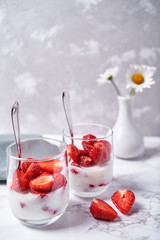 Two glasses of healthy yogurt with fresh sliced strawberry, spoons and chamomile flowers in vase on white table background, copy space. Healthy breakfast, food concept