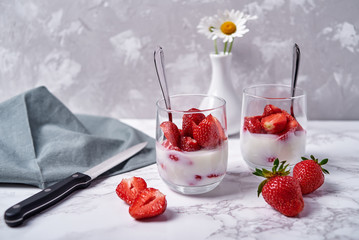 Two glasses of healthy yogurt with fresh sliced strawberry, spoons and chamomile flowers in vase on...