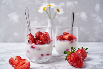 Two glasses of healthy yogurt with fresh sliced strawberry, spoons and chamomile flowers in vase on white table background, copy space. Healthy breakfast, food concept