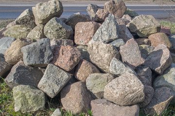 A pile of granite boulders with jagged sharp edges in gray, beige, and brown colors piled on top of each other on the roadside grass. Mineral material for decorative construction and cladding.