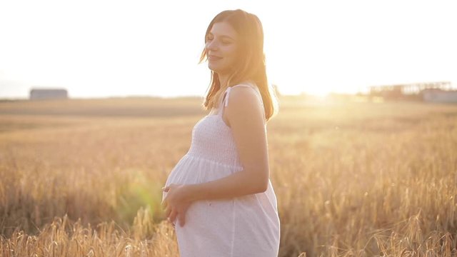 Stock footage of an attractive pregnant woman in white summer dress running in the gold field of rye in bright sunlight in the evening.