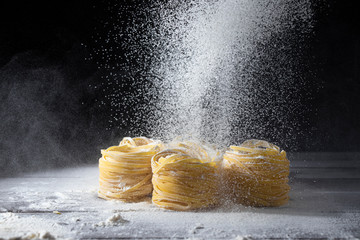 Flour is sifted through a sieve on Raw tagliatelle pasta on a wooden kitchen table. Black background