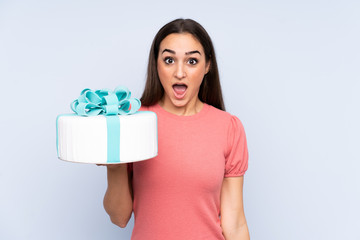 Pastry chef holding a big cake isolated on blue background with surprise and shocked facial expression