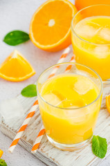 Freshly squeezed orange juice with ice in a glass with a straw on a wooden board on a light background with fresh oranges. Vertical orientation, close up.