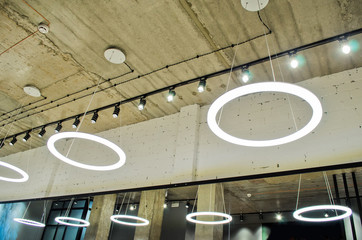 White circular ring shaped lamps hang under a concrete ceiling with external wiring and black...