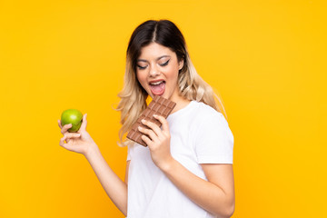 Teenager girl isolated on  yellow background taking a chocolate tablet in one hand and an apple in...