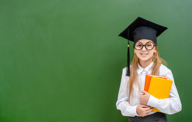 Smiling girl wearing funny eyeglasses and a graduation cap holds books near a school board. Empty space for text