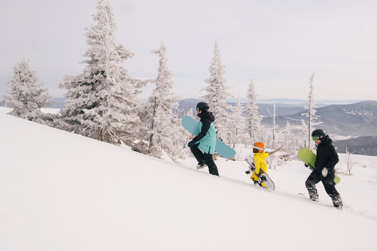 Family of snowboarders walking on mountain top. Sunny holiday in ski resort, snow covered spurs trees, mountains landscape