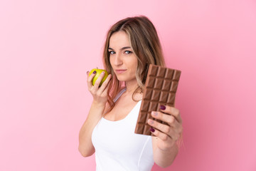 Young woman over isolated pink background taking a chocolate tablet in one hand and an apple in the...