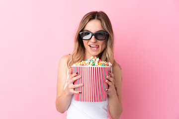 Young woman over isolated pink background with 3d glasses and holding a big bucket of popcorns