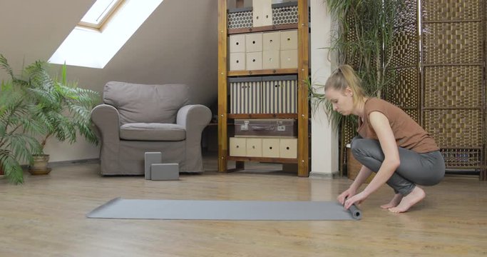 Blonde woman rolls up her yoga mat in her home studio.