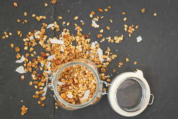 Top view of a glass jar up to the top filled with organic granola with nuts and coconut. Jar with muesli on a black stone background. Healthy lifestyle concept.