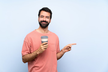 Young man with beard holding a take away coffee over isolated blue background pointing finger to the side