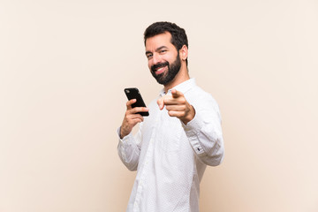 Young man with beard holding a mobile points finger at you with a confident expression