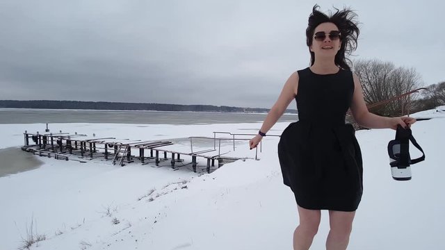 Woman in black dress and sunglasses jumps and dances on river bank covered with snow to make herself warm. Front view. Copy space.
