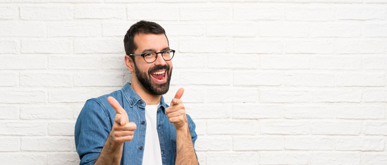 Handsome man with beard over white brick wall pointing to the front and smiling