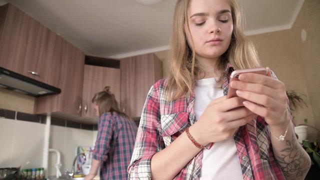 Young married couple in the kitchen. A girl with a tattoo writes a message on her mobile phone to her lover on the background of her husband who washes the dishes.