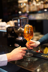 Person hands an orange aperol cocktail to another person. Classical italian aperitif. Alcoholic beverage with lots of ice. Vertical close up photo. Selective focus on glass, blurred background.