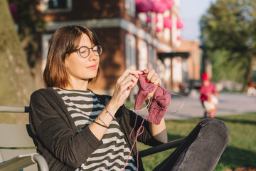 Obraz premium Close up photo of cute pretty girl in glasses and striped t-shirt. Attractive woman sitting in chair and knitting socks from purple yarn outdoor. Needleworks and hobby concept.