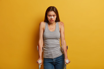 Serious unhappy woman beaten by cruel people, suffers from various fractures, poses with mobility aid equipment against yellow background. Female biker knocked off bicycle by vehicle, tries to recover