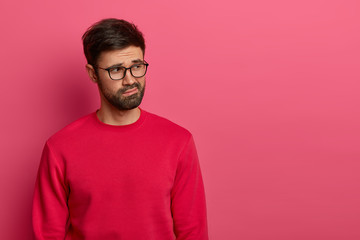 Photo of thoughtful puzzled man with dissatisfied expression concentrated aside, tries to recall information, has thick beard, wears spectacles and red sweater, looks unhappily on blank space