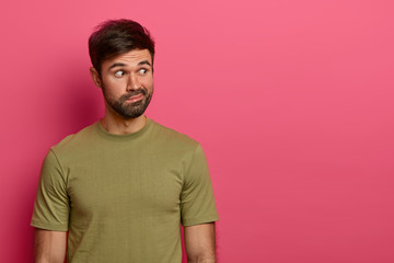 Obraz premium Photo of surprised bearded guy concentrated aside with shocked expression, thinks deeply about something, purses lips, wears casual olive t shirt, makes decision, poses against rosy background