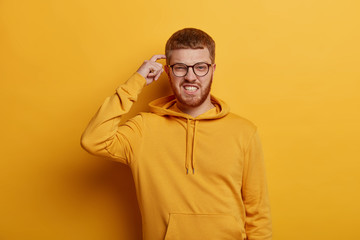 Obraz premium Confused bearded red head man scratches head, squints face and clenches teeth, has bad memory, tries to recall something wears casual sweatshirt isolated on yellow wall plans or makes difficult choice