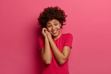 Tender pleasant looking woman keeps hands near face, has positive happy look, dressed in casual red t shirt, concentrated on pleasant thoughts, isolated on pink background, rejoices spare time