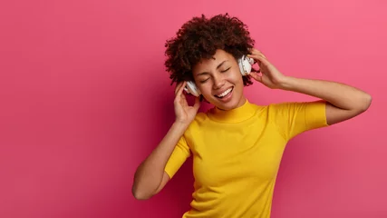 Poster Overjoyed African American woman listens modern music via wireless headphones, enjoys good sound and chills with eyes closed, smiles broadly, wears casual yellow t shirt, isolated on pink wall © Wayhome Studio