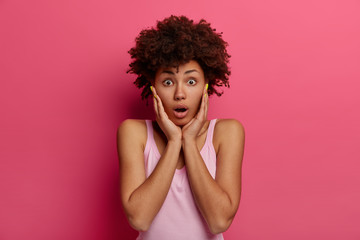 Horizontal shot of stupefied emotional African American woman touches cheeks with hands, stares at camera with disbelief, gasps from fear and wonder, dressed casually, poses indoor. Human reaction