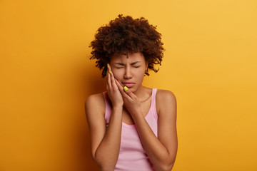 Displeased dark skinned woman suffers from toothache, touches cheek, needs to see dentist, take painkillers, has health problems, closes eyes with pain, dressed casually, isolated on yellow background