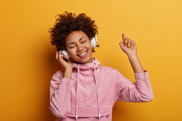 Happy female teenager enjoys pleasant music, wears stereo headphones, dances with arm raised, feels upbeat, catches every tune of song, wears rosy velvet sweatshirt, isolated on yellow background