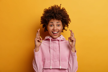 Obraz na płótnie Canvas Positive dark skinned young woman with Afro hairstyle looks hopefully at camera, awaits admission university, believes in good luck, waits for something, wears hoodie, poses over yellow background