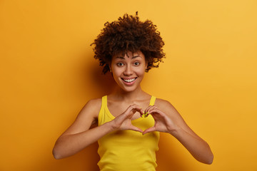 Isolated shot of pleasant looking woman makes heart gesture, says be my valentine, smiles positively, expresses love and care, fell in love with someone, wears yellow shirt, stands indoor alone.