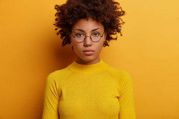 Headshot of serious looking woman with Afro hairstyle, looks directly at camera, wears round spectacles and yellow jumper, thinks over about something important, stands in studio. Face expression