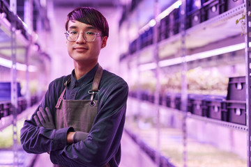 Portrait of Asian young farmer in apron standing with arms crossed and looking at camera while working in the farm