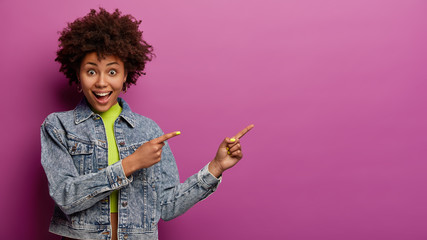 Positive surprised African American woman points aside at blank space, has glad expression,...