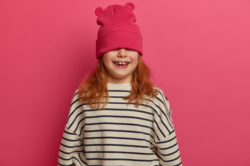 Carefree adorable girl plays indoor, enjoys great day, expresses positive attitude, hides face with hat, wears loose jumper with black stripes, poses against pink background. Children, fun concept